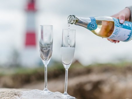 2 Gläser auf einem Stein, in diese "Geltinger Bucht Sekt" eingegossen wird mit Leuchtturm im Hintergrund