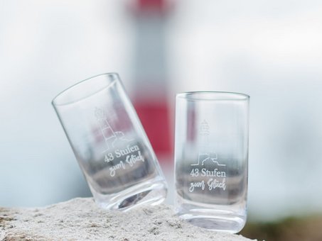 2 Gläser mit Schriftzug "43 Stufen zum Glück" auf einem Stein mit Leuchtturm im Hintergrund